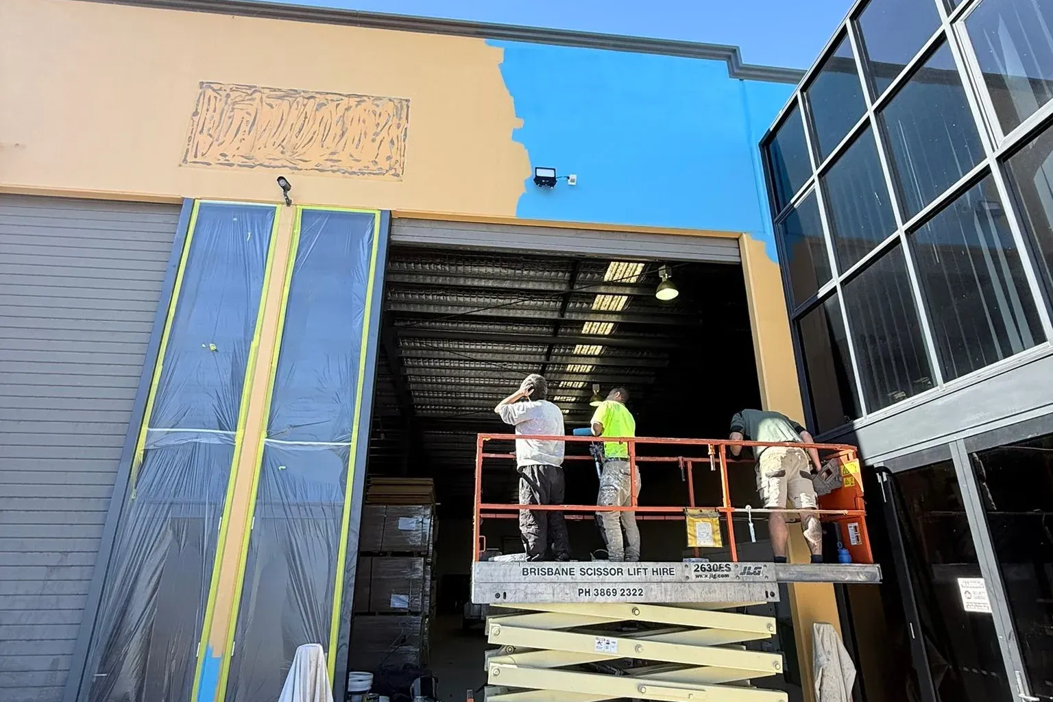 AnB painters on a scissor lift repainting a commercial warehouse facade in Rothwell, half still orange and half finished in bright blue